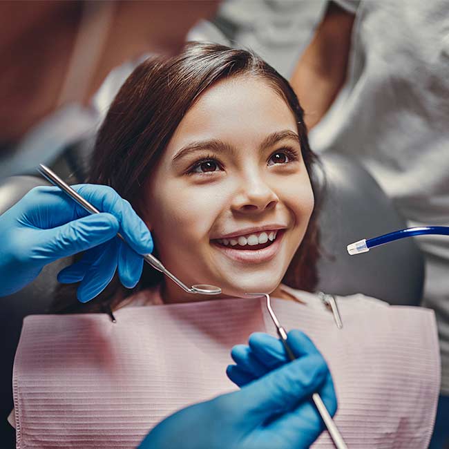 A young girl smiles as a dental progressional with blue glove prepares to use dental tools