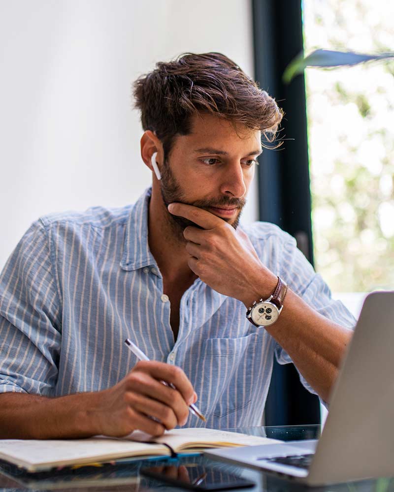 A man listens to ear pods while taking notes as he looks at a laptop computer