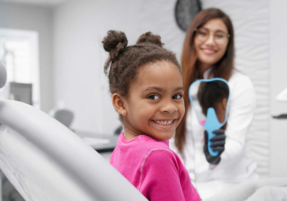 Little african girl sitting on dentist chair looking at camera and smiling while female dentist keeping mirror at backround