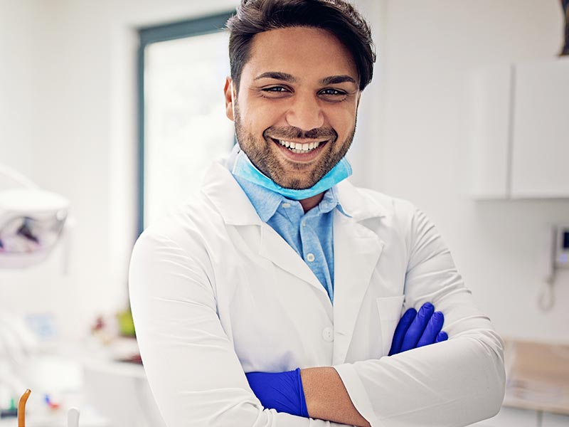 Smiling man in white lab coat and gloves looks at the camera - he's most likely a dentist